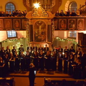 großer Chor singend in einer Kirche. Im Hintergrund der beleuchtete Weihnachtsbaum