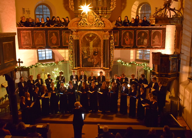 großer Chor singend in einer Kirche. Im Hintergrund der beleuchtete Weihnachtsbaum